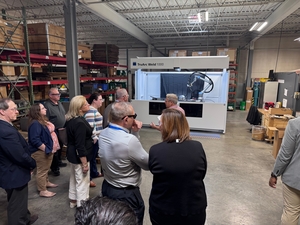 Group of people in a warehouse gathered around a large white industrial CNC machine with robotic arm and safety enclosure.