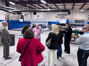 Group of people standing in a factory or print shop, listening to a man in a pink shirt near large industrial machines.