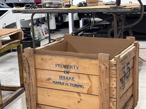 Wooden industrial crate marked "PROPERTY OF HDAKE MANUFACTURING" with stenciled numbers, sitting in a workshop.