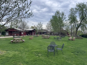Open grassy park with scattered picnic tables and metal chairs, trees and low wooden buildings under a cloudy sky