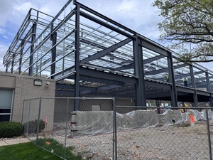 Steel building framework rising above a low existing structure, fenced construction site with gravel and protective sheeting.