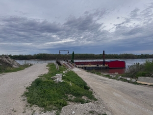 Gravel road splitting toward a riverbank with a red barge moored at a small dock under a cloudy sky, low vegetation on the embankment.