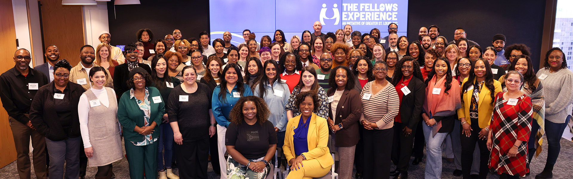 Large diverse group of smiling adults standing and seated in an office meeting room wearing name tags, with a screen behind them reading "The Fellows Experience."