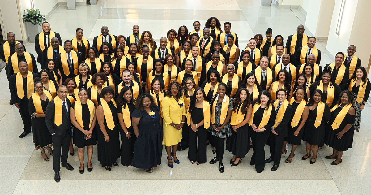 Large diverse group of adults dressed in black with golden yellow stoles, standing in rows in a bright atrium for a formal group portrait.