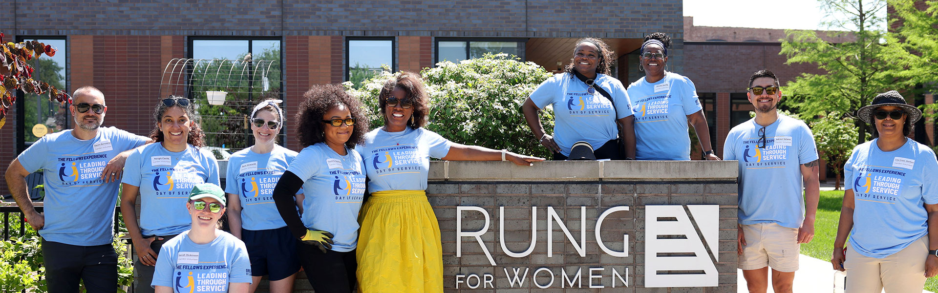 Diverse group of volunteers in matching blue "Leading Through Service" shirts smiling behind a RUNG for Women sign outdoors
