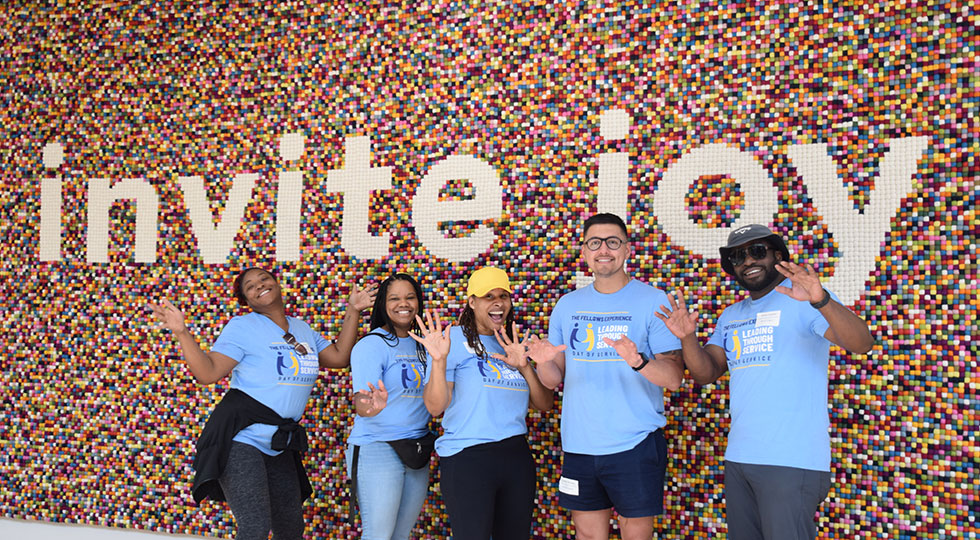 Five smiling people in light blue event shirts waving in front of a colorful mosaic wall spelling "invite joy."