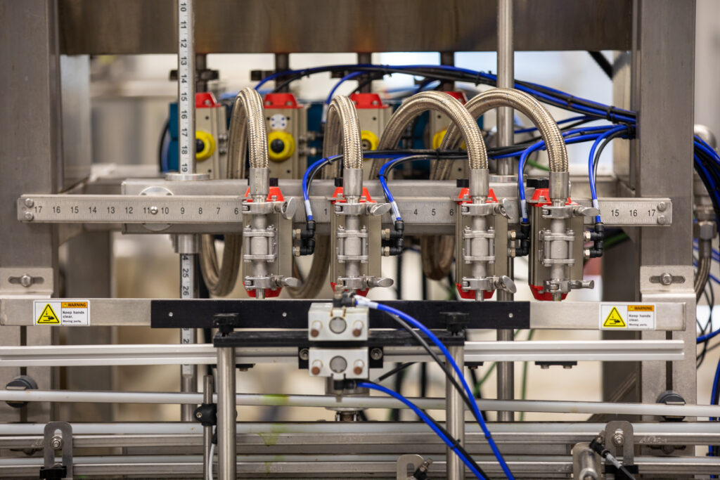 Close-up of a four-nozzle liquid filling machine with braided steel hoses, blue air lines, and warning labels