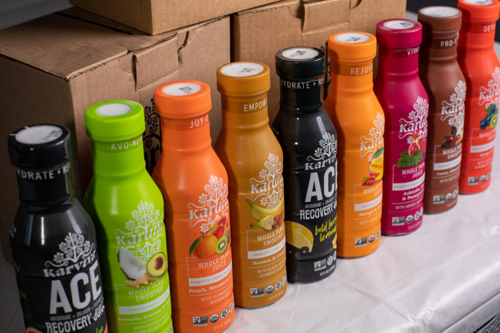 Row of colorful Karina juice and smoothie bottles in various flavors lined up in front of cardboard boxes
