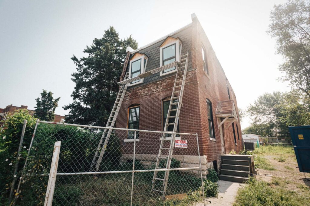 Two-story brick house under renovation with ladders leaning against it, chain-link rental fence, and a dumpster nearby