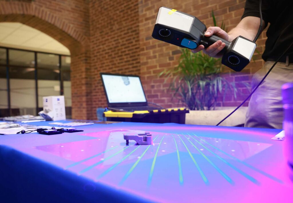 Person uses a handheld 3D laser scanner projecting green lines onto a small object, with a laptop showing the scan