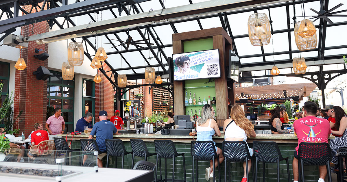 Patrons sit at a bustling open-air bar with woven pendant lights, glass roof, and a screen advertising a Surf Mesa event
