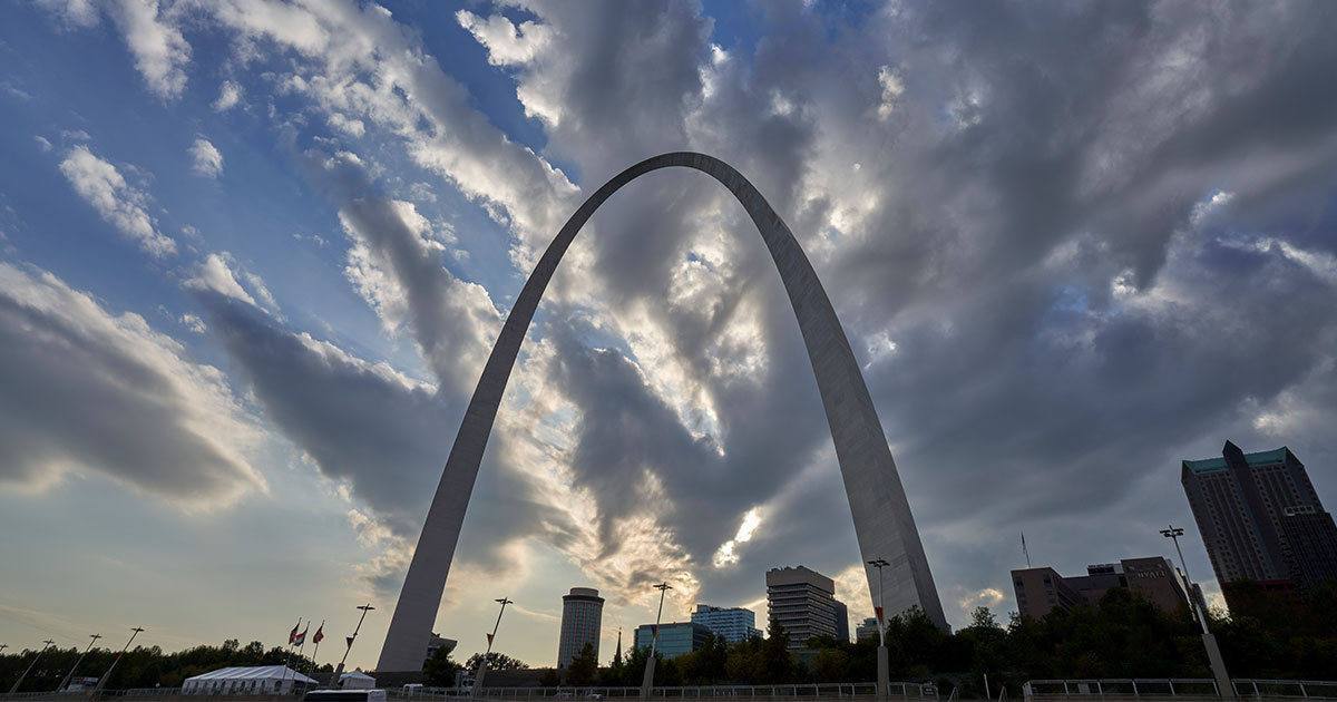 The Gateway Arch in St. Louis rises above the city skyline under a dramatic cloudy sky