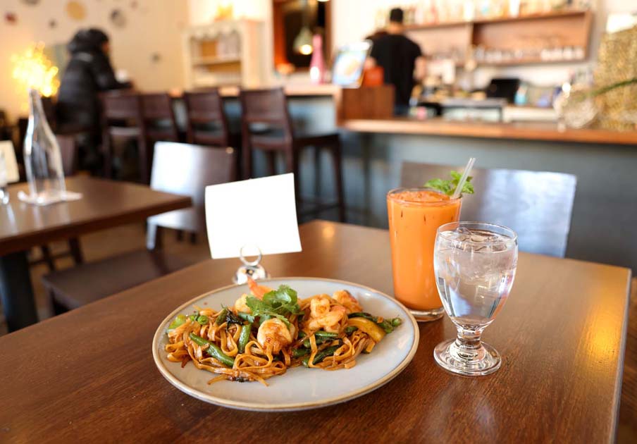 Aubergine Cafe 5 Shrimp stir-fried noodles, iced Thai tea, and water on a wooden restaurant table