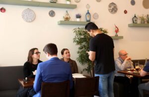 A server takes an order from three seated diners in a restaurant with decorative wall plates and shelves