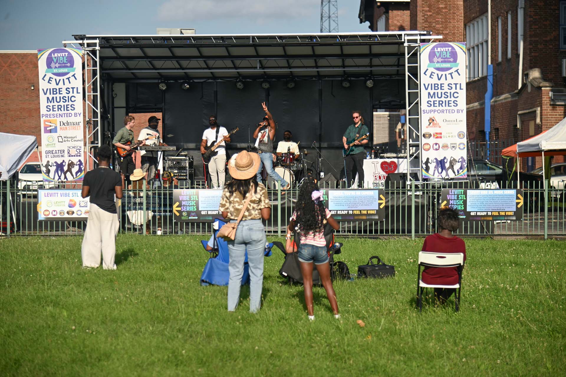 DELMAR FESTIVAL MT 2026 015 A band performs on the Levitt Vibe STL Music Series stage as a small crowd watches from a grassy lawn