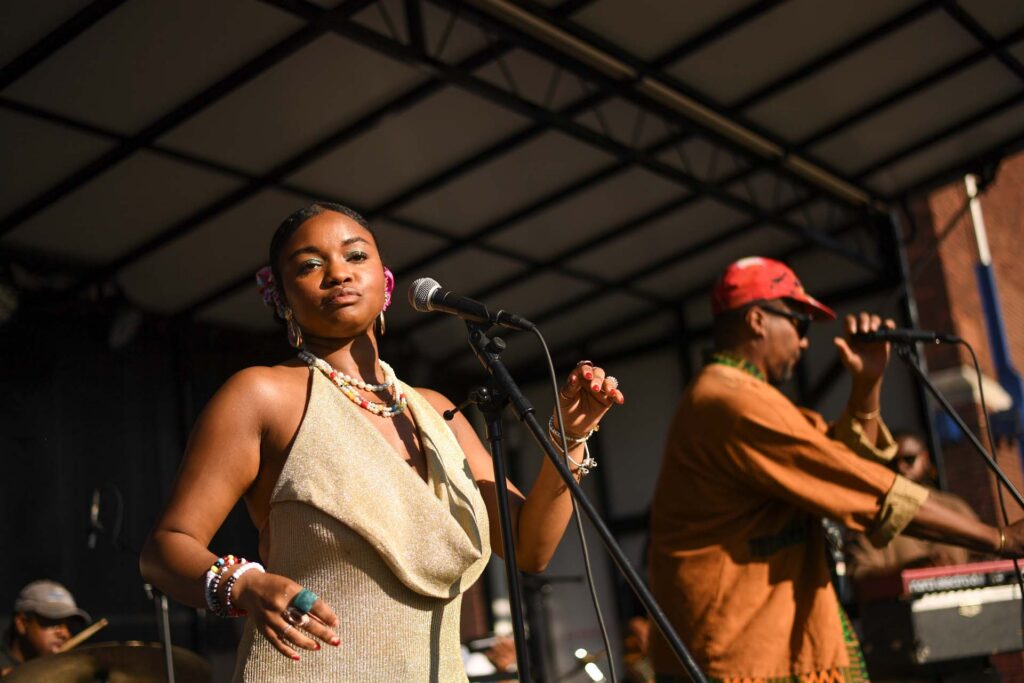 Woman in gold dress and beaded jewelry sings at a microphone on an outdoor stage beside a male vocalist in a red cap