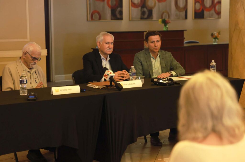 Three men sit at a panel table with name cards and microphones addressing an audience member