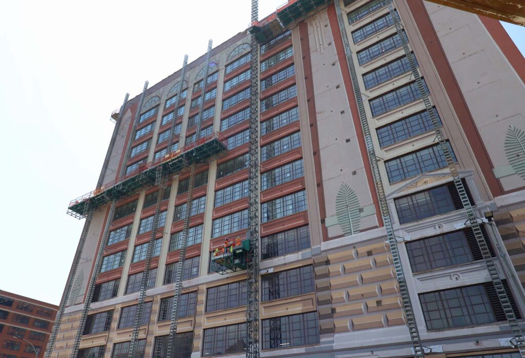 Workers on a suspended platform work on the ornate facade of a large high-rise building under renovation
