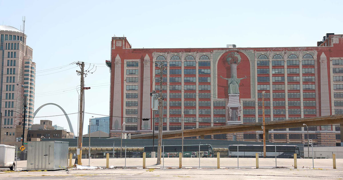 Large red building with a mural of a robed figure, near St. Louis Gateway Arch and power lines