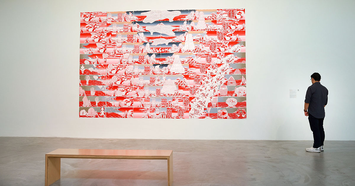 A person views a large red and white detailed artwork on a gallery wall, with a wooden bench nearby