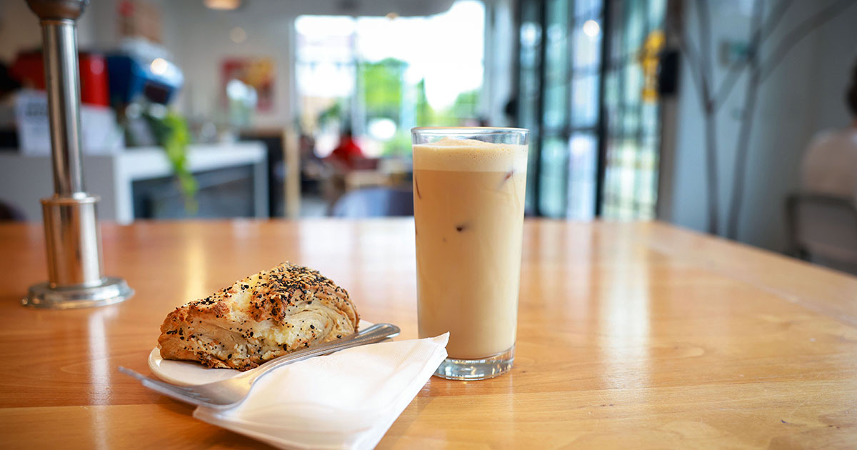 Iced latte in a tall glass beside a seeded pastry on a plate at a sunlit cafe table