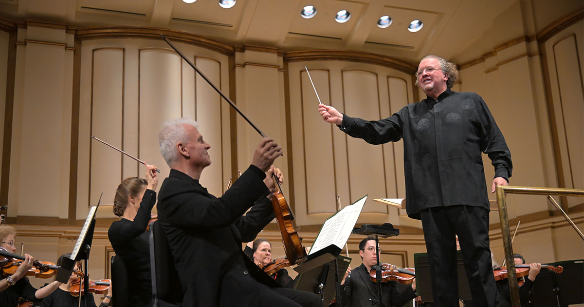 Conductor with raised baton leads an orchestra of string players on a concert hall stage