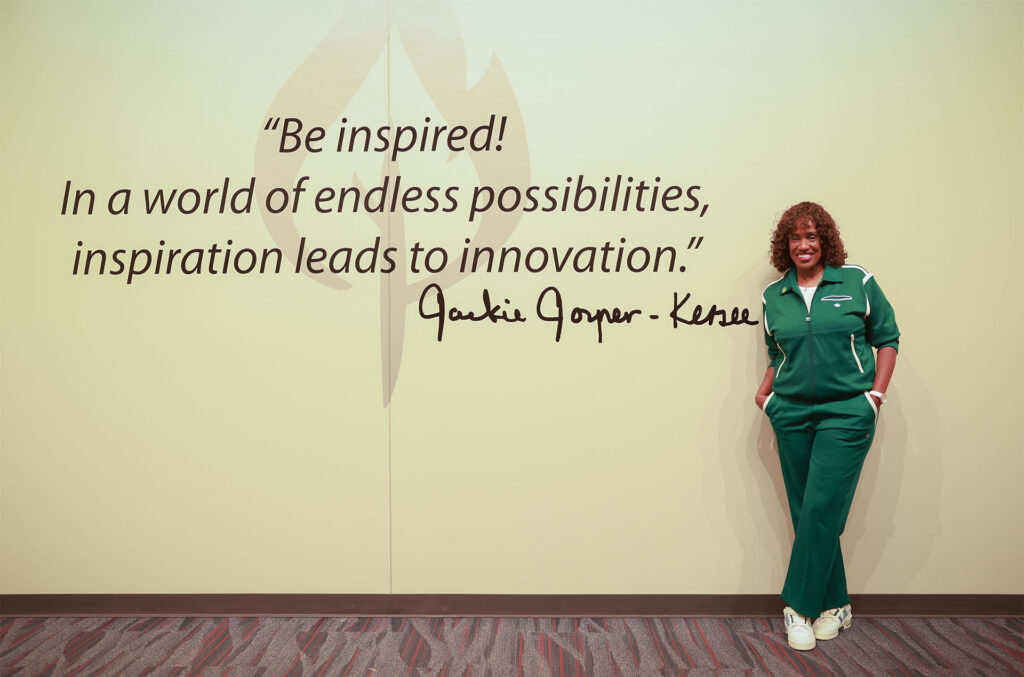 Smiling woman in a green tracksuit leans against a wall with a Jackie Joyner-Kersee quote about inspiration leading to innovation