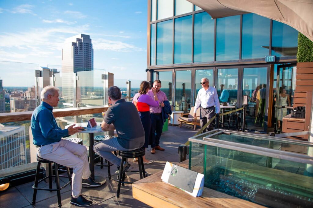 People socializing at a sunny rooftop bar with glass railings overlooking a city skyline