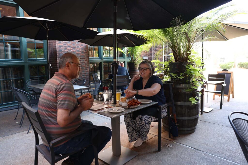 A man and smiling woman share a meal at an outdoor restaurant patio under black umbrellas beside potted palms