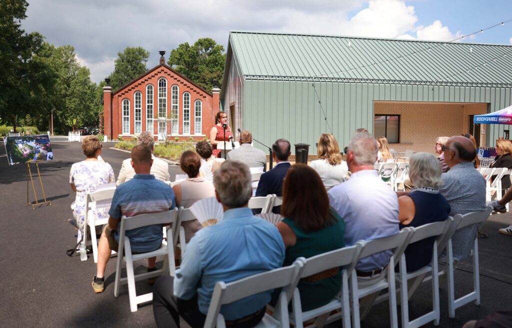 Woman in polka-dot dress speaks at an outdoor event to a seated audience near a historic brick building