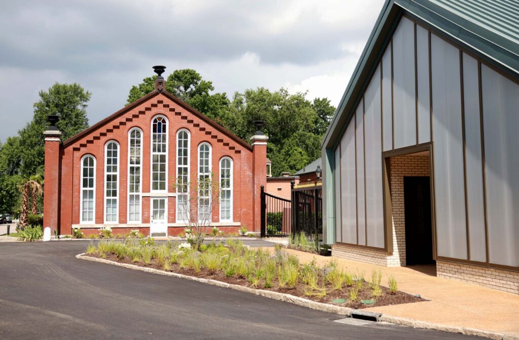 Historic red brick building with tall arched windows beside a modern metal-clad structure, framed by landscaped gardens under a cloudy sky