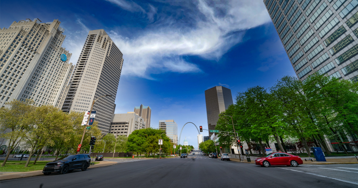 Downtown St. Louis street framed by skyscrapers and trees, with the Gateway Arch visible under a blue sky
