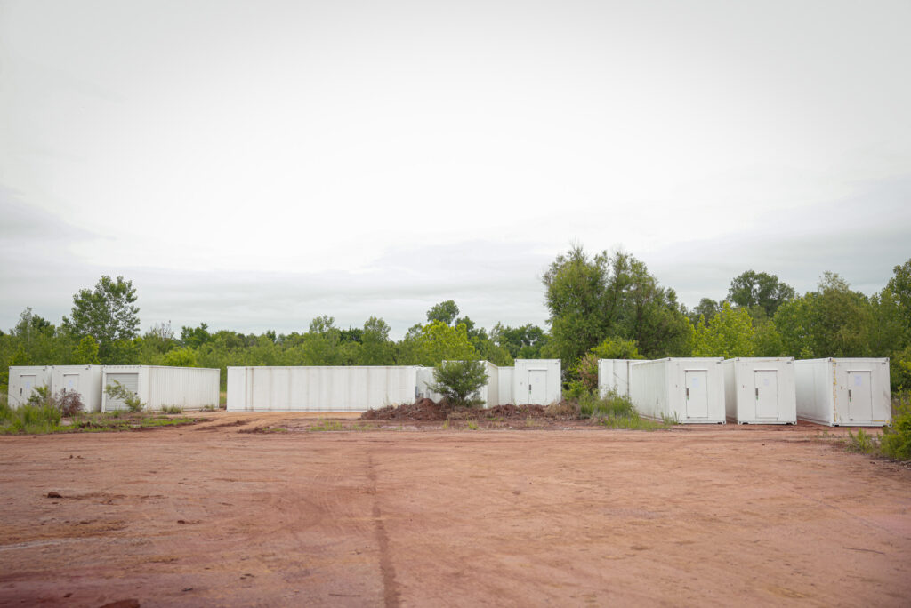 Row of white shipping containers on a red dirt lot backed by green trees under an overcast sky
