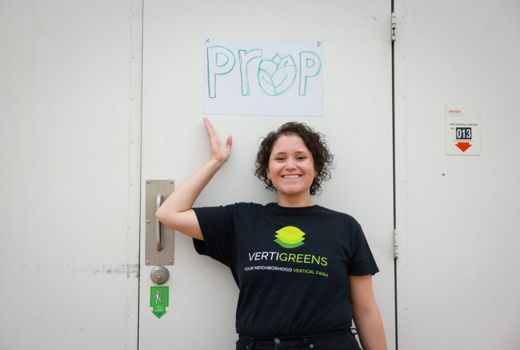 Smiling woman in a black VertiGreens vertical farm shirt gestures toward a hand-drawn Prop sign on a white door