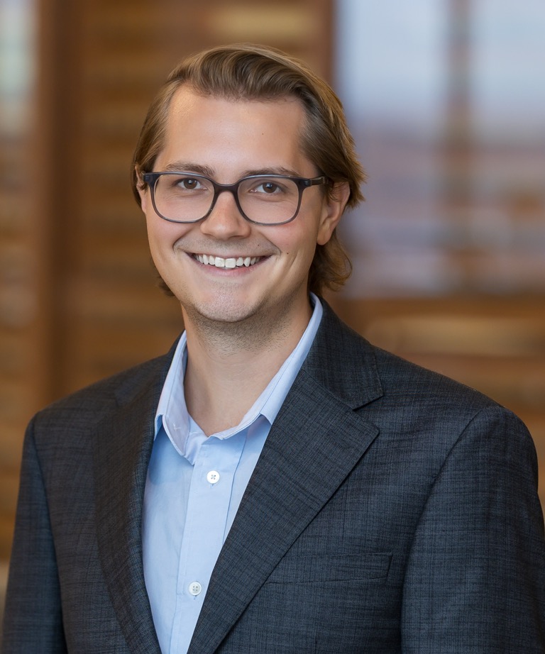 Smiling man with glasses wearing a dark blazer and light blue shirt in a professional headshot