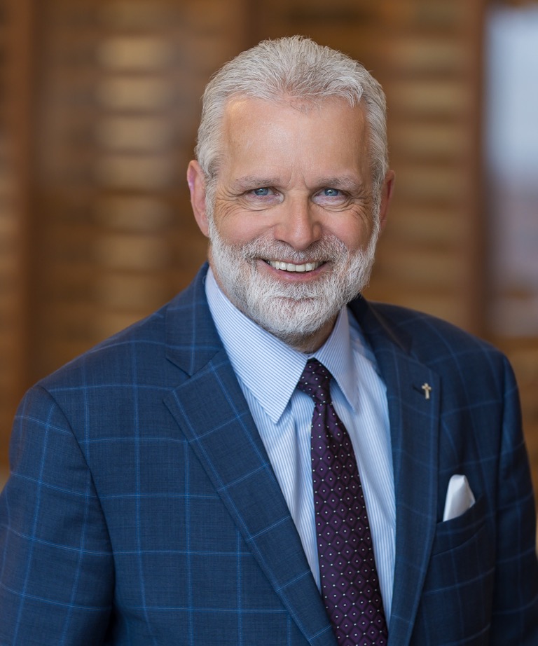 Smiling man with white beard in a blue plaid suit and purple tie, wearing a cross lapel pin