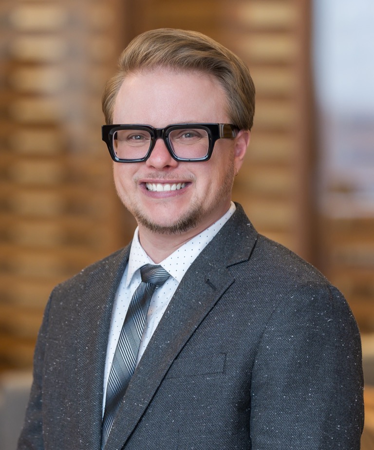 Smiling man with black-framed glasses and beard wearing a gray suit and striped tie in a professional headshot