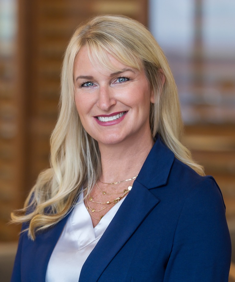 Smiling blonde woman in a navy blazer and white top, professional headshot with blurred background