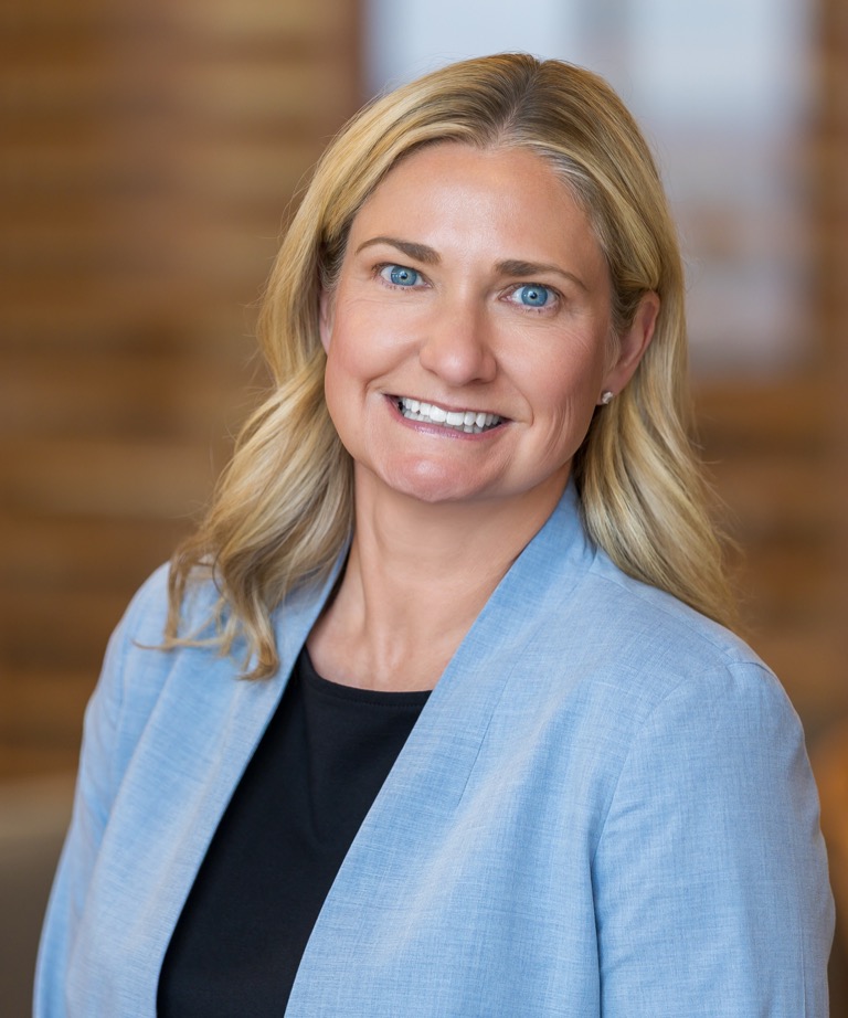 Smiling blond woman in a light blue blazer and black top, professional headshot with blurred warm background
