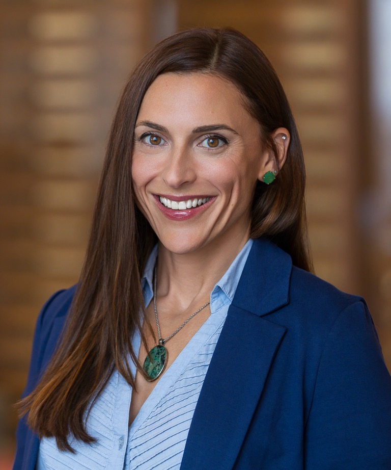 Smiling woman with long brown hair wearing a navy blazer, light blue shirt, and green pendant necklace