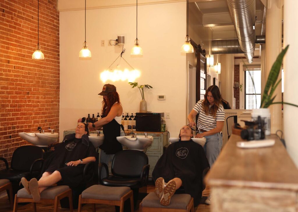 Two stylists wash clients' hair at wash basins in a salon with exposed brick walls and pendant lighting