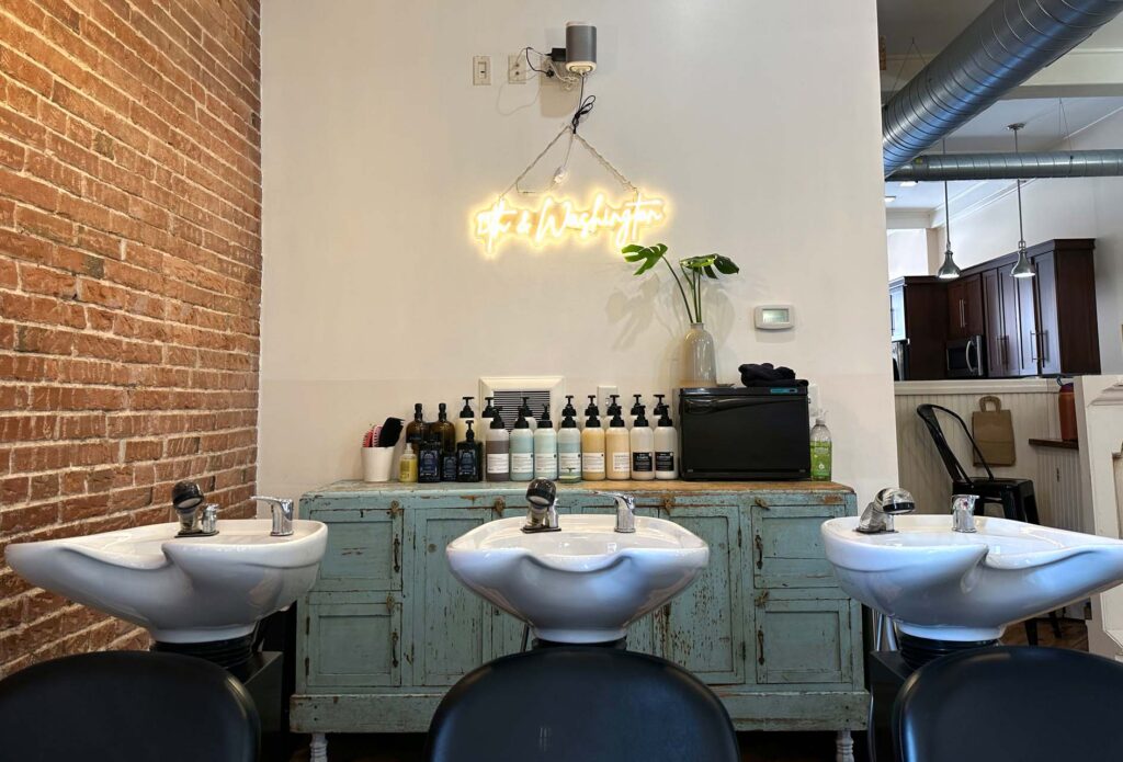 Salon hair-washing station with three sinks, product bottles on a vintage cabinet, and a glowing neon script sign