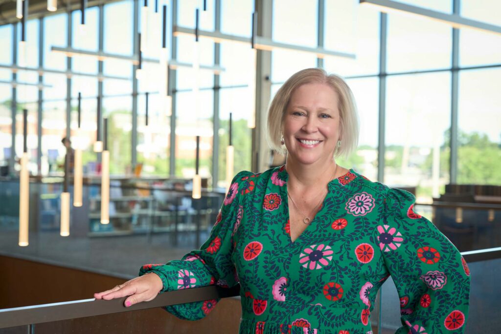Smiling woman in a green floral dress leaning on a railing in a bright, glass-walled interior