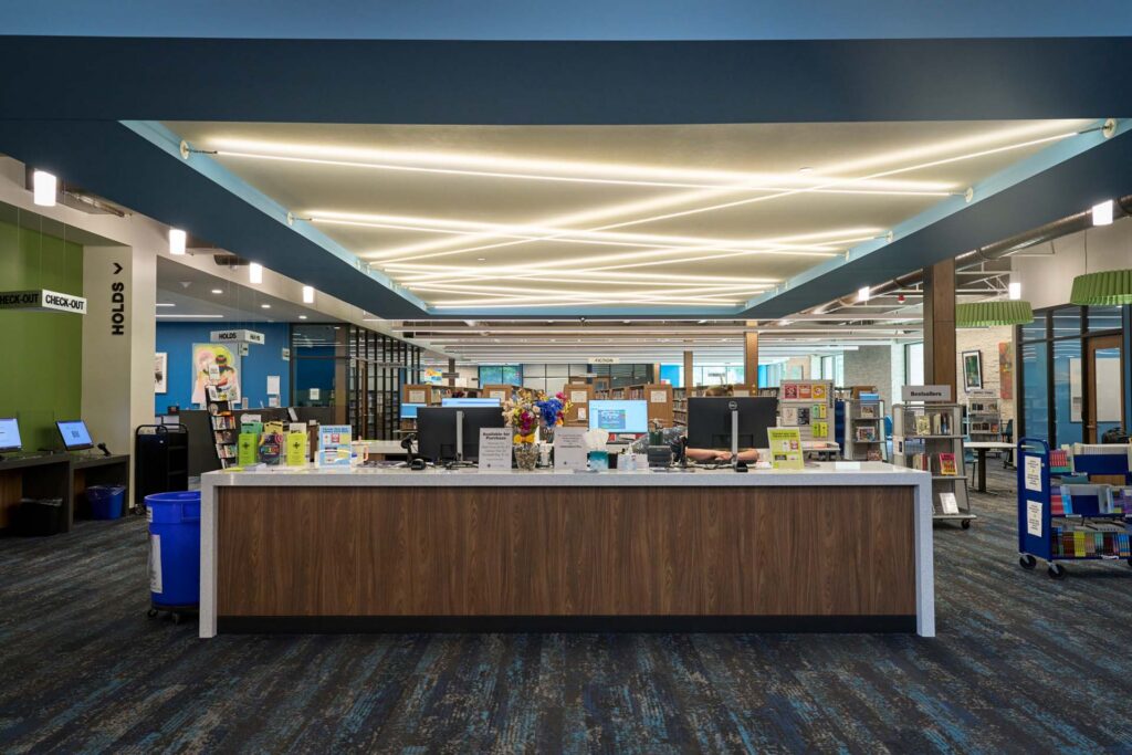 Library service desk with computers under angular ceiling lighting, with check-out, holds, and book shelving areas nearby