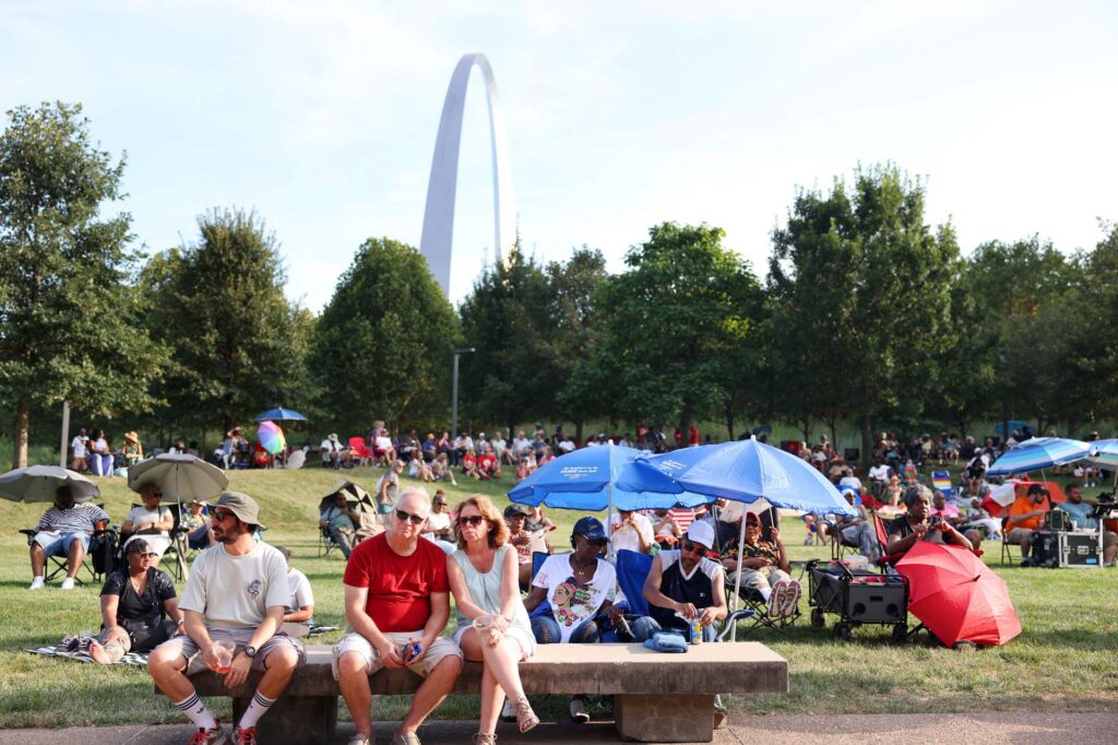 Crowds relax on lawn chairs and benches under umbrellas at an outdoor event near the Gateway Arch in St. Louis