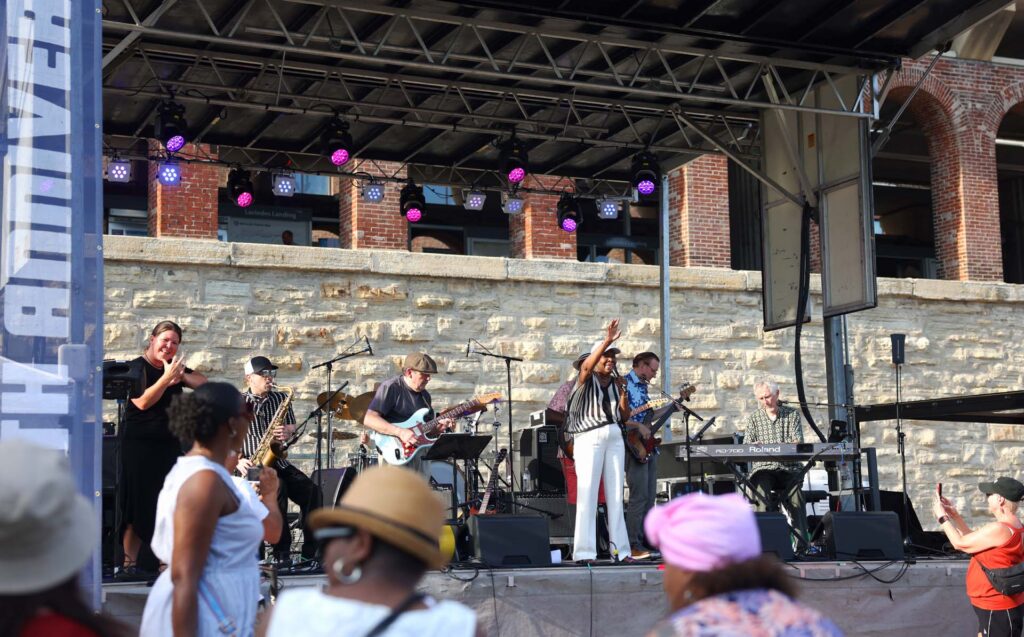 Band performs on an outdoor stage with a singer, saxophonist, guitarists, and keyboardist as a crowd watches