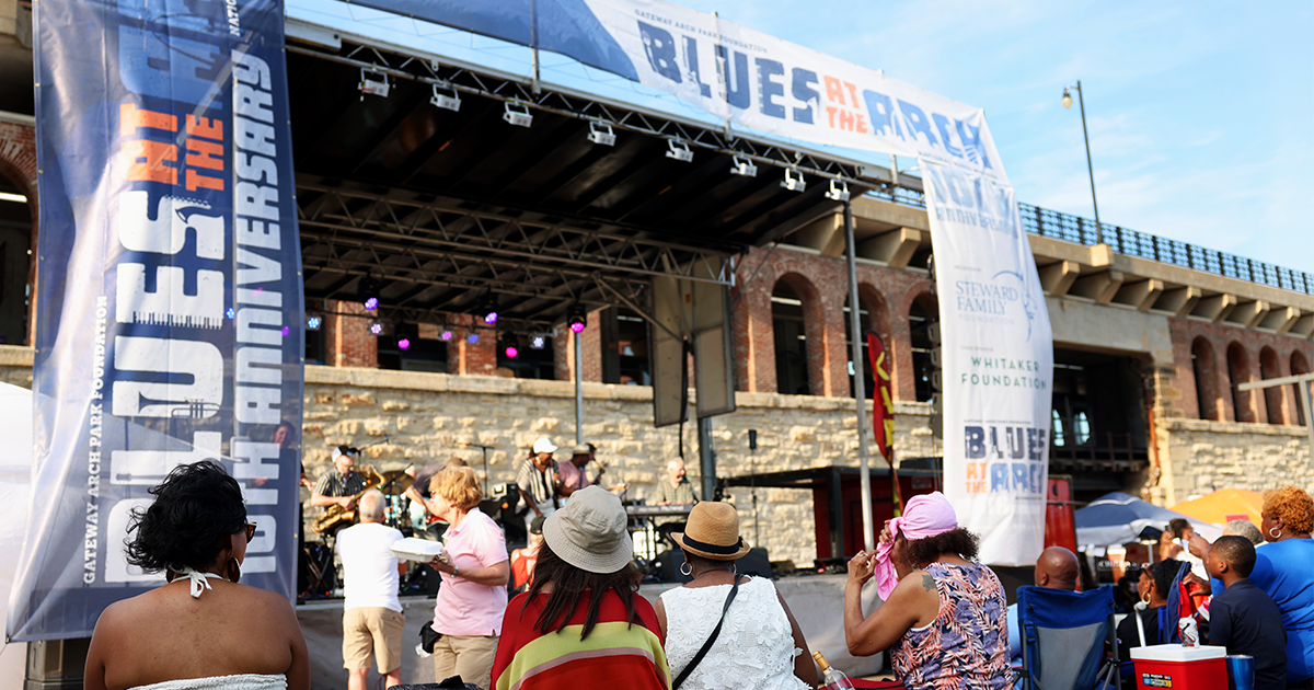 Blues at the Arch featured photo Crowd watches musicians perform at outdoor Blues at the Arch festival stage with banners on a sunny day