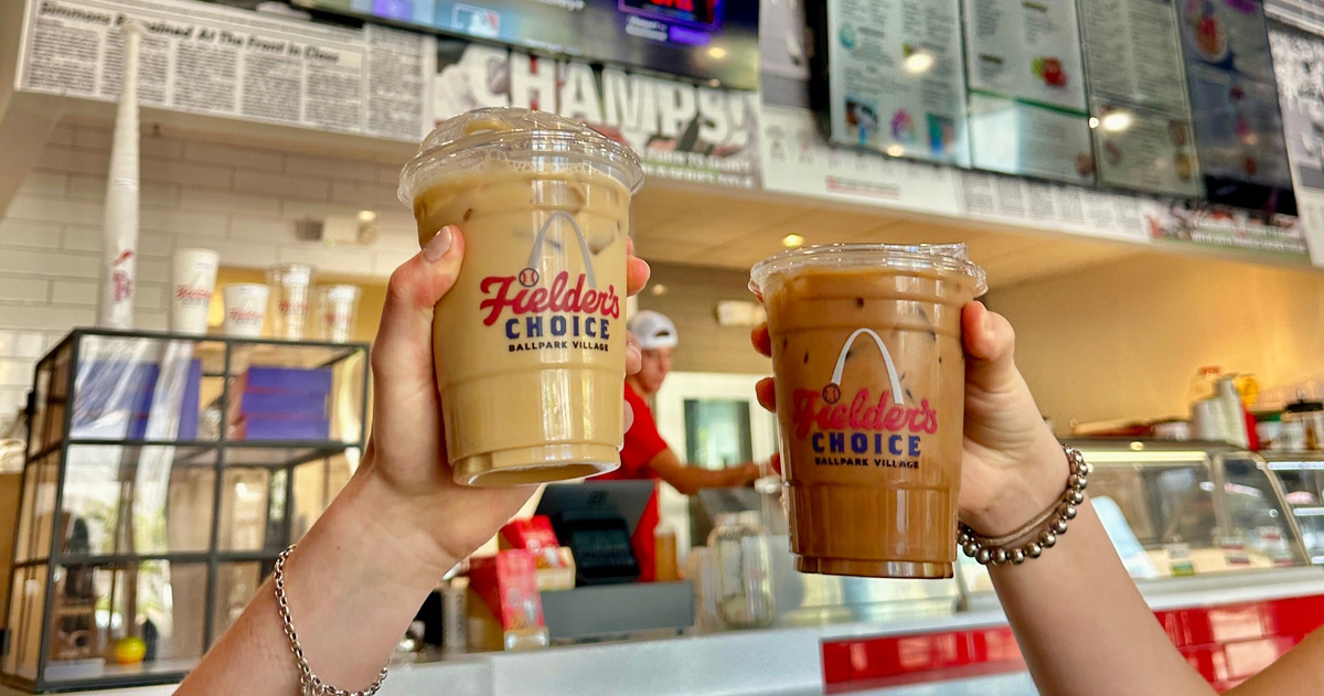 Two hands hold up iced coffees in Fielder's Choice Ballpark Village cups at a cafe counter