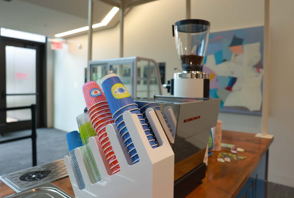 Coffee shop counter with colorful Goshen-branded cups in a dispenser, a coffee grinder, and an espresso machine