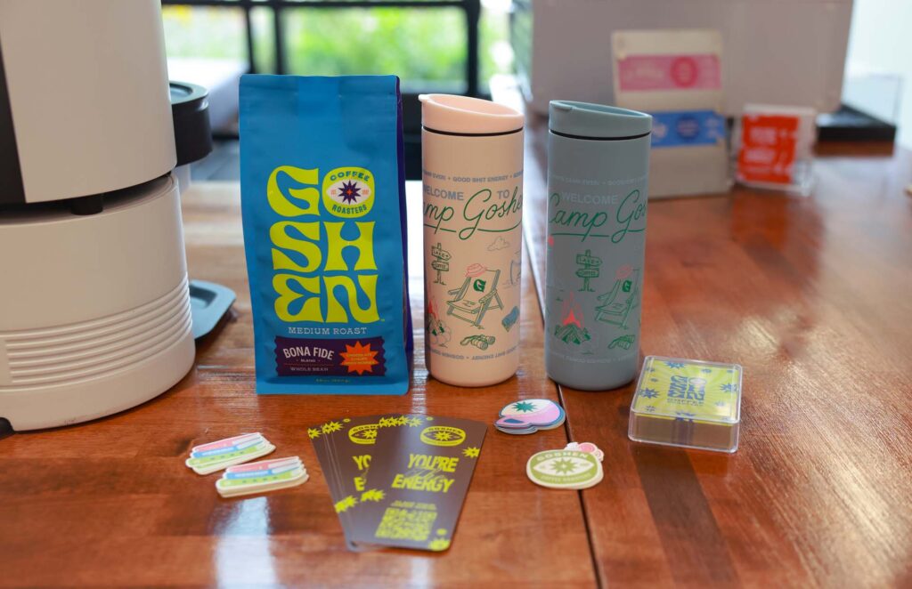 Goshen Coffee Roasters bag of Bona Fide medium roast, two "Camp Goshen" travel tumblers, stickers, and cards on a wooden table
